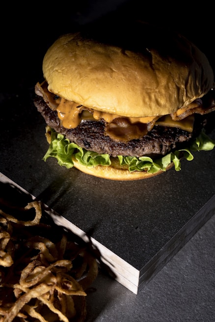 A close-up of a juicy burger with a soft toasted bun, melted cheese, crispy bacon, lettuce, and a generous drizzle of sauce. The burger is placed on a slate surface alongside a pile of golden onion rings, illuminated by dramatic lighting.