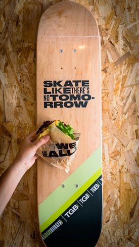 A skateboard with a natural wood finish features the phrase 'Skate Like There's No Tomorrow' in bold black text. A hand is holding a wrapped burger over the skateboard. The setting appears to be against a textured wooden surface, adding a rustic feel.