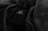 A sleek black perfume bottle lies on a smooth, dark fabric. The bottle has a minimalist design with subtle reflections on its surface, giving it a luxurious and refined appearance.