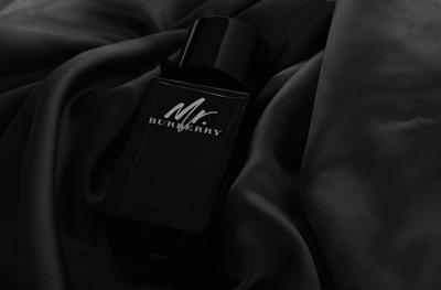 A sleek black perfume bottle lies on a smooth, dark fabric. The bottle has a minimalist design with subtle reflections on its surface, giving it a luxurious and refined appearance.