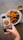 A hand is holding a smartphone, capturing a photo of crispy fried food served in a white bowl lined with branded paper. There is a small dipping sauce container next to the bowl.