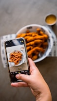A hand is holding a smartphone, capturing a photo of crispy fried food served in a white bowl lined with branded paper. There is a small dipping sauce container next to the bowl.