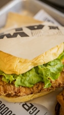 A close-up of a fried chicken sandwich with crisp lettuce and a soft bun, wrapped partially in branded wax paper. The sandwich sits on a tray lined with paper featuring printed text.