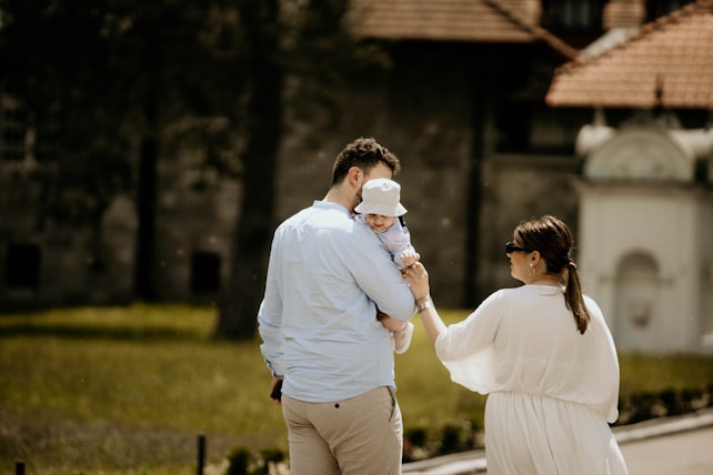 A family of three is enjoying a walk outdoors, with a man carrying a baby who is wearing a sun hat. A woman walks beside them, holding the man's arm. They appear to be in a park or garden area with a blurred building in the background.