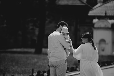 A family scene with two adults and a baby captured in a tender moment. The adults, a woman and a man, are walking outdoors, with the man carrying the baby. The setting appears to be a sunny day with some historical architecture in the background. The image is in black and white, enhancing the nostalgic and classic feel.