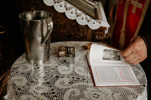 A finely crocheted lace tablecloth covers a round table. On the table, there is a large metal vessel, resembling a pitcher, and an open book with a black-and-white image on one of its pages. The person is wearing a garment with red and gold colors, appearing ceremonial or religious. In the background, there is ornate wooden furniture partially visible.