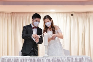 A couple wearing formal attire and face masks are participating in a candle lighting ceremony. The man is dressed in a black suit with a bow tie, and the woman is wearing a white gown. They both hold candles, lighting a central candle together on a table covered with a white cloth. The background consists of beige curtains.