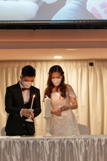 A man and a woman are wearing formal attire and face masks while lighting candles together on a table. The setting suggests a ceremonious or wedding atmosphere. The backdrop features a white curtain, and a large lit candle stands between them as they each light smaller candles.