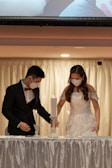 A couple dressed in formal attire, wearing face masks, stand together behind a table adorned with white fabric and candles. The scene suggests a formal event or ceremony with a soft and intimate atmosphere.