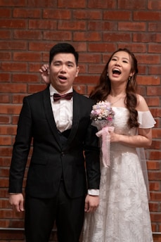 A couple dressed in formal attire stands in front of a brick wall. The woman is wearing a white wedding gown and holding a bouquet of flowers, smiling joyfully. The man is in a dark suit with a red bow tie, displaying a surprised expression.
