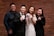A group of four people are posed in front of a brick wall. Two are wearing formal attire, with one woman in a white dress holding a bouquet of flowers and a man wearing a black suit with a bow tie. Two others are casually dressed, with one in a denim jacket and the other in a black outfit. They appear to be smiling and gesturing with their hands.