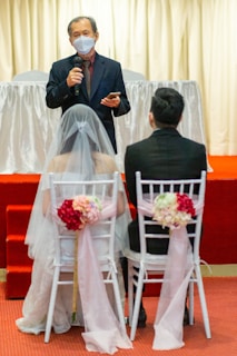 A wedding ceremony scene with a person speaking while holding a microphone, standing in front of a bride and groom seated on white chairs adorned with pink flowers and ribbons. The speaker is wearing a mask and a suit, set against a backdrop with light curtains and a red carpet.