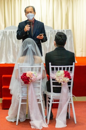 A wedding ceremony scene with a person speaking while holding a microphone, standing in front of a bride and groom seated on white chairs adorned with pink flowers and ribbons. The speaker is wearing a mask and a suit, set against a backdrop with light curtains and a red carpet.