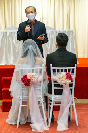A wedding ceremony scene with a person speaking while holding a microphone, standing in front of a bride and groom seated on white chairs adorned with pink flowers and ribbons. The speaker is wearing a mask and a suit, set against a backdrop with light curtains and a red carpet.