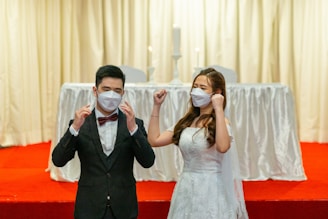 A couple dressed in formal wedding attire is standing on a red carpeted platform. The bride is wearing a white gown and the groom is in a black suit with a red bow tie. Both are wearing white face masks. The background features a white draped table with two tall white candles.