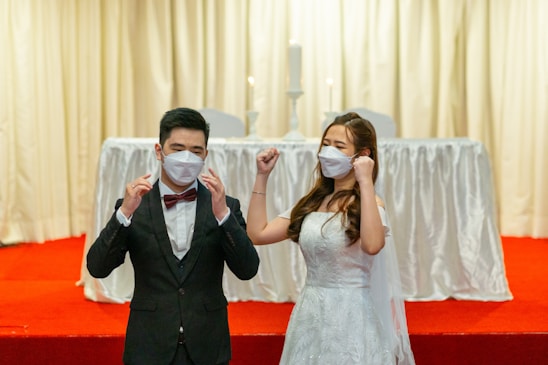 A couple dressed in formal wedding attire is standing on a red carpeted platform. The bride is wearing a white gown and the groom is in a black suit with a red bow tie. Both are wearing white face masks. The background features a white draped table with two tall white candles.