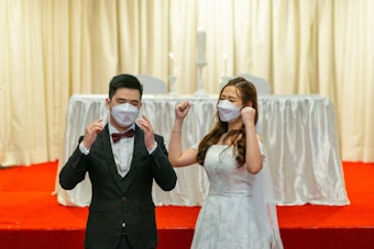 A couple dressed in formal wedding attire is standing on a red carpeted platform. The bride is wearing a white gown and the groom is in a black suit with a red bow tie. Both are wearing white face masks. The background features a white draped table with two tall white candles.