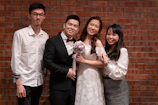 Four people are posing in front of a brick wall. One is wearing a wedding dress and holding a bouquet, while the others are dressed in formal and semi-formal attire.