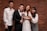 Four people are posing in front of a brick wall. One is wearing a wedding dress and holding a bouquet, while the others are dressed in formal and semi-formal attire.