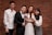 Four people are posing in front of a brick wall. One is wearing a wedding dress and holding a bouquet, while the others are dressed in formal and semi-formal attire.