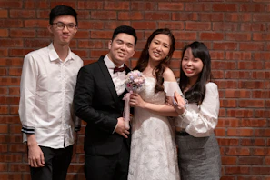 Four people are posing in front of a brick wall. One is wearing a wedding dress and holding a bouquet, while the others are dressed in formal and semi-formal attire.