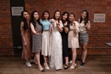 Seven women are standing in front of a brick wall, smiling and making various hand gestures. The woman in the center is wearing a white dress and holding a bouquet, while the others are dressed in various casual and semi-formal attire. There are signs on the wall behind them that say Thank you for clearing your own plates and Please maintain social distancing. Do not join the tables.