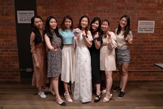Seven women are standing in front of a brick wall, smiling and making various hand gestures. The woman in the center is wearing a white dress and holding a bouquet, while the others are dressed in various casual and semi-formal attire. There are signs on the wall behind them that say Thank you for clearing your own plates and Please maintain social distancing. Do not join the tables.