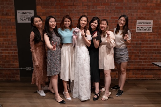 Seven women are standing in front of a brick wall, smiling and making various hand gestures. The woman in the center is wearing a white dress and holding a bouquet, while the others are dressed in various casual and semi-formal attire. There are signs on the wall behind them that say Thank you for clearing your own plates and Please maintain social distancing. Do not join the tables.