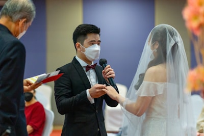 A couple is exchanging vows during a wedding ceremony. The groom, wearing a black suit and a bow tie, is speaking into a microphone while holding the bride's hand. The bride is dressed in a white gown and veil. Both are wearing face masks. An officiant is seen on the left, holding a book and wearing a mask.