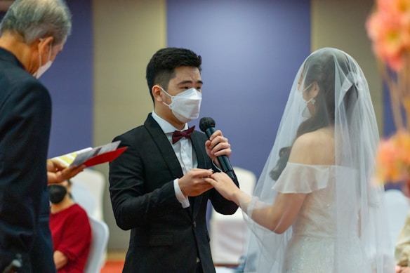 A couple is exchanging vows during a wedding ceremony. The groom, wearing a black suit and a bow tie, is speaking into a microphone while holding the bride's hand. The bride is dressed in a white gown and veil. Both are wearing face masks. An officiant is seen on the left, holding a book and wearing a mask.