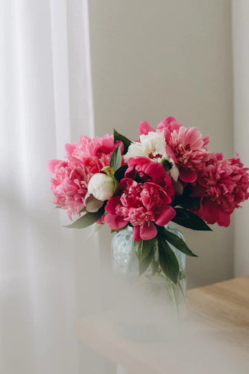 A close-up of a delicate floral arrangement featuring soft pink peonies and white ranunculus in a sleek glass vase.