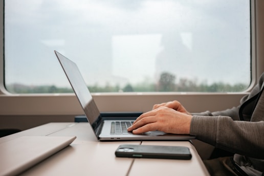 The interior of a train cabin with a person typing on a laptop placed on a table next to a smartphone. The window reveals a blurred view of the outdoors, suggesting movement. The person is casually dressed in a hoodie, focusing on the laptop.