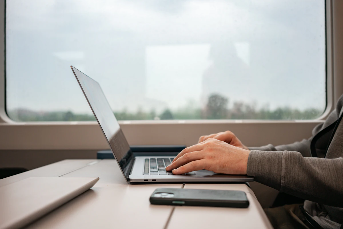 Hands type on an open laptop at a train table by the window, a phone lying beside it and green countryside blurred outside