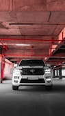 A white SUV is parked in an industrial-style parking garage, characterized by exposed concrete surfaces and red pipes that form geometric patterns on the ceiling. The vehicle is centered with headlights on, casting a distinct shadow on the concrete floor.