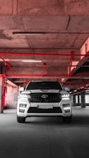 A white SUV is parked in an industrial-style parking garage, characterized by exposed concrete surfaces and red pipes that form geometric patterns on the ceiling. The vehicle is centered with headlights on, casting a distinct shadow on the concrete floor.