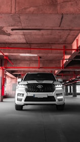 A white SUV is parked in an industrial-style parking garage, characterized by exposed concrete surfaces and red pipes that form geometric patterns on the ceiling. The vehicle is centered with headlights on, casting a distinct shadow on the concrete floor.