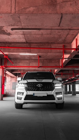 A white SUV is parked in an industrial-style parking garage, characterized by exposed concrete surfaces and red pipes that form geometric patterns on the ceiling. The vehicle is centered with headlights on, casting a distinct shadow on the concrete floor.