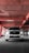 A white SUV is parked in an industrial-style parking garage, characterized by exposed concrete surfaces and red pipes that form geometric patterns on the ceiling. The vehicle is centered with headlights on, casting a distinct shadow on the concrete floor.