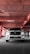 A white SUV is parked in an industrial-style parking garage, characterized by exposed concrete surfaces and red pipes that form geometric patterns on the ceiling. The vehicle is centered with headlights on, casting a distinct shadow on the concrete floor.