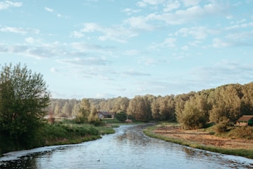 A serene river winds through a lush green landscape bordered by dense trees. A small house is nestled among the foliage, and the sky overhead is a soft, cloudy blue. The peaceful setting suggests a quiet countryside scene.