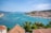 A scenic coastal view with turquoise waters, lined with boats and yachts along the marina. A hill covered with buildings having terracotta roofs is visible in the background, and lush greenery including palm trees is in the foreground.