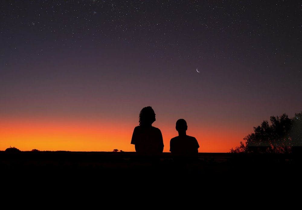 a couple of people sitting on top of a hill under a night sky