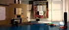 An open electrical panel with a multitude of colorful wires connected to various circuit breakers is mounted on a wall. Tools are placed on a blue workbench, including a multimeter and a screwdriver. A visible checklist is attached to the left panel door.