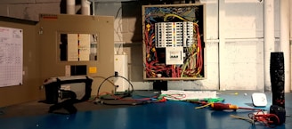 An open electrical panel with a multitude of colorful wires connected to various circuit breakers is mounted on a wall. Tools are placed on a blue workbench, including a multimeter and a screwdriver. A visible checklist is attached to the left panel door.