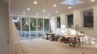 A large, bright room under construction with several large windows and glass doors revealing an outdoor scene with trees and patio furniture. Inside, there are tools, saws, and wooden planks scattered across two workbenches. The walls are freshly painted white, and recessed ceiling lights are illuminated, adding to the spacious and airy feel.