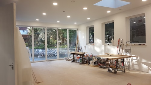 A large, bright room under construction with several large windows and glass doors revealing an outdoor scene with trees and patio furniture. Inside, there are tools, saws, and wooden planks scattered across two workbenches. The walls are freshly painted white, and recessed ceiling lights are illuminated, adding to the spacious and airy feel.