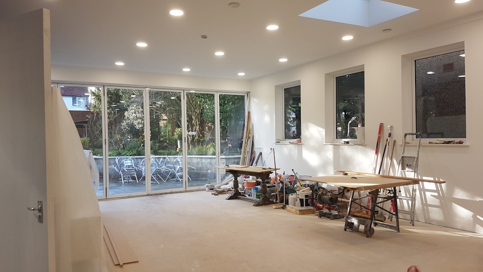 A large, bright room under construction with several large windows and glass doors revealing an outdoor scene with trees and patio furniture. Inside, there are tools, saws, and wooden planks scattered across two workbenches. The walls are freshly painted white, and recessed ceiling lights are illuminated, adding to the spacious and airy feel.