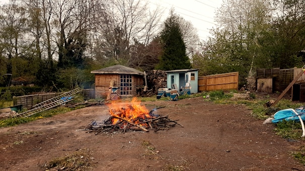 A small outdoor area featuring a central bonfire surrounded by a few wooden structures. There's a log cabin style shed with a window and a small blue shed nearby, both enveloped by dense greenery. Various tools and pieces of equipment, like a ladder and a wheelbarrow, are scattered around, with a wooden fence running along the right. The environment appears to be a rustic garden or backyard.
