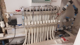 A complex manifold system with multiple white pipes connected in parallel to a row of valves. Several gauges are visible, as well as an assortment of knobs and control switches. The setup appears part of a heating or plumbing system, with wiring and connectors visible. The background shows a partially unfinished wall, suggesting an installation in progress or maintenance area.