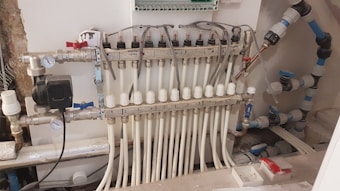 A complex manifold system with multiple white pipes connected in parallel to a row of valves. Several gauges are visible, as well as an assortment of knobs and control switches. The setup appears part of a heating or plumbing system, with wiring and connectors visible. The background shows a partially unfinished wall, suggesting an installation in progress or maintenance area.
